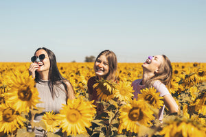 Three women, with colorful Nöz sunscreen on their noses, laughing in a field of giant sunflowers against a blue sky